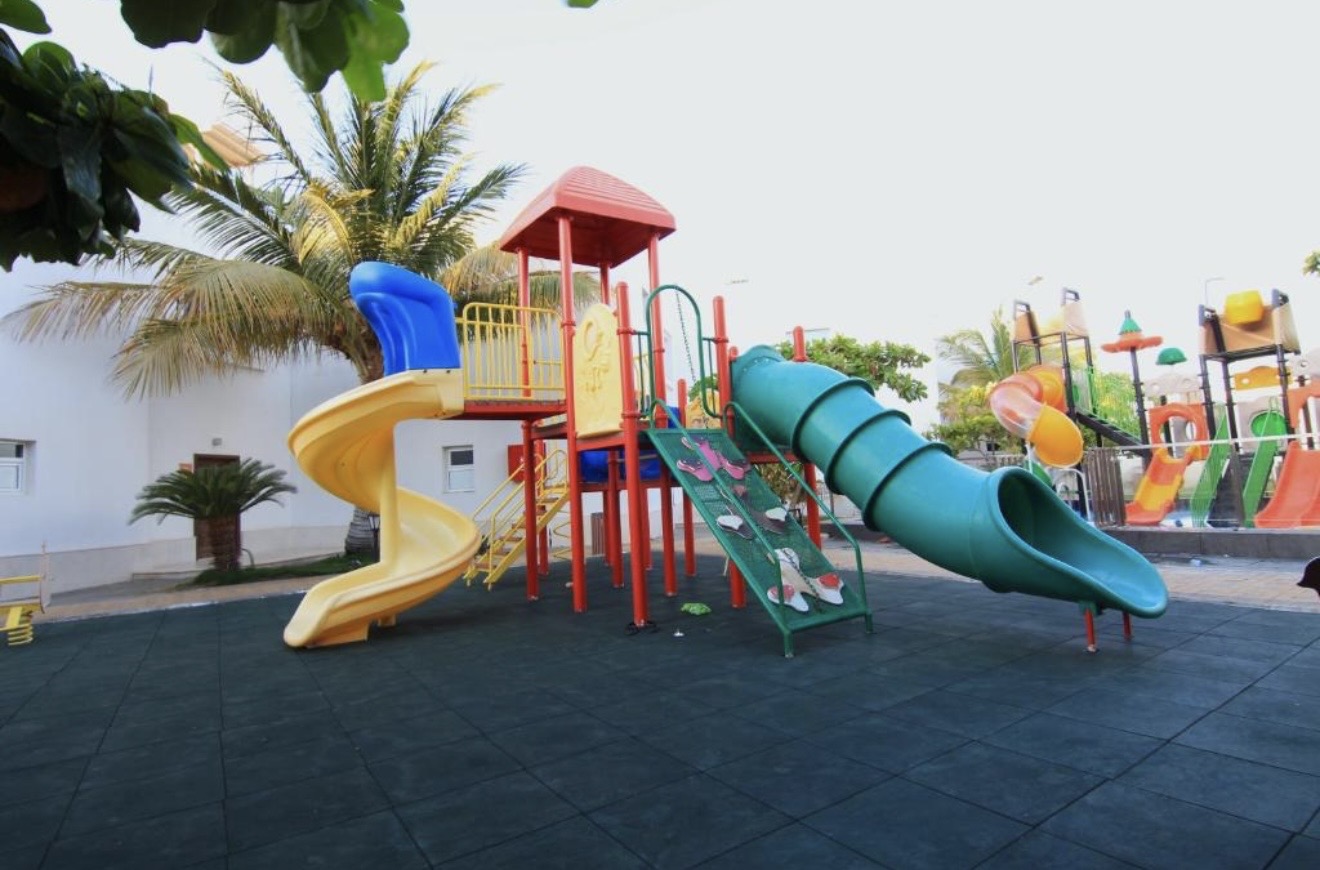 ملعب أطفال بأرضية مطاطية فيه زحلقة حلزونية صفراء وأنبوب أخضر، والألعاب المائية خلفه.